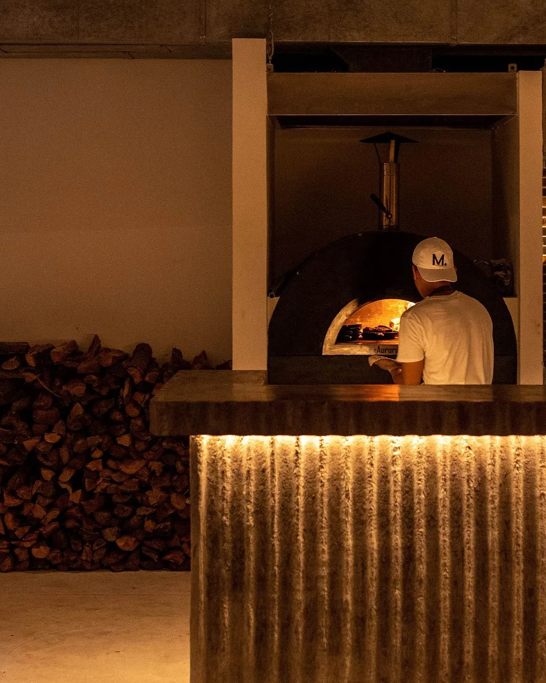 Person wearing a white cap and shirt placing or removing food from a wood-fired oven in a dimly lit setting with stacked firewood nearby.