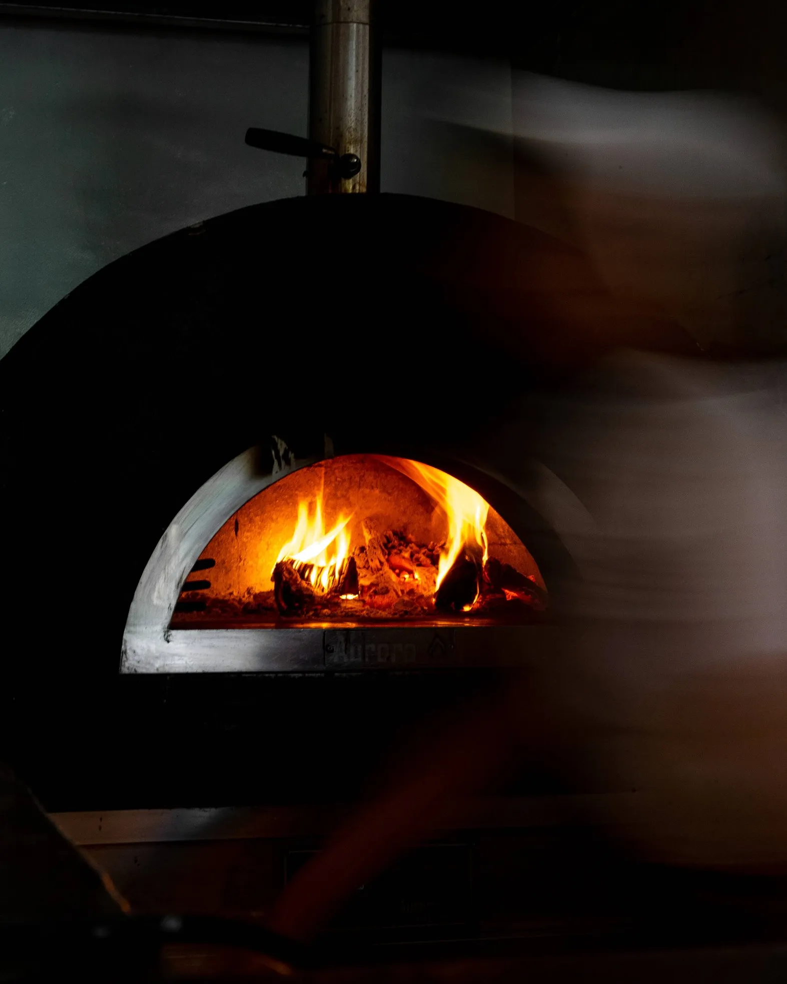 Wood-fired pizza oven with bright flames inside and blurred motion of a person nearby.