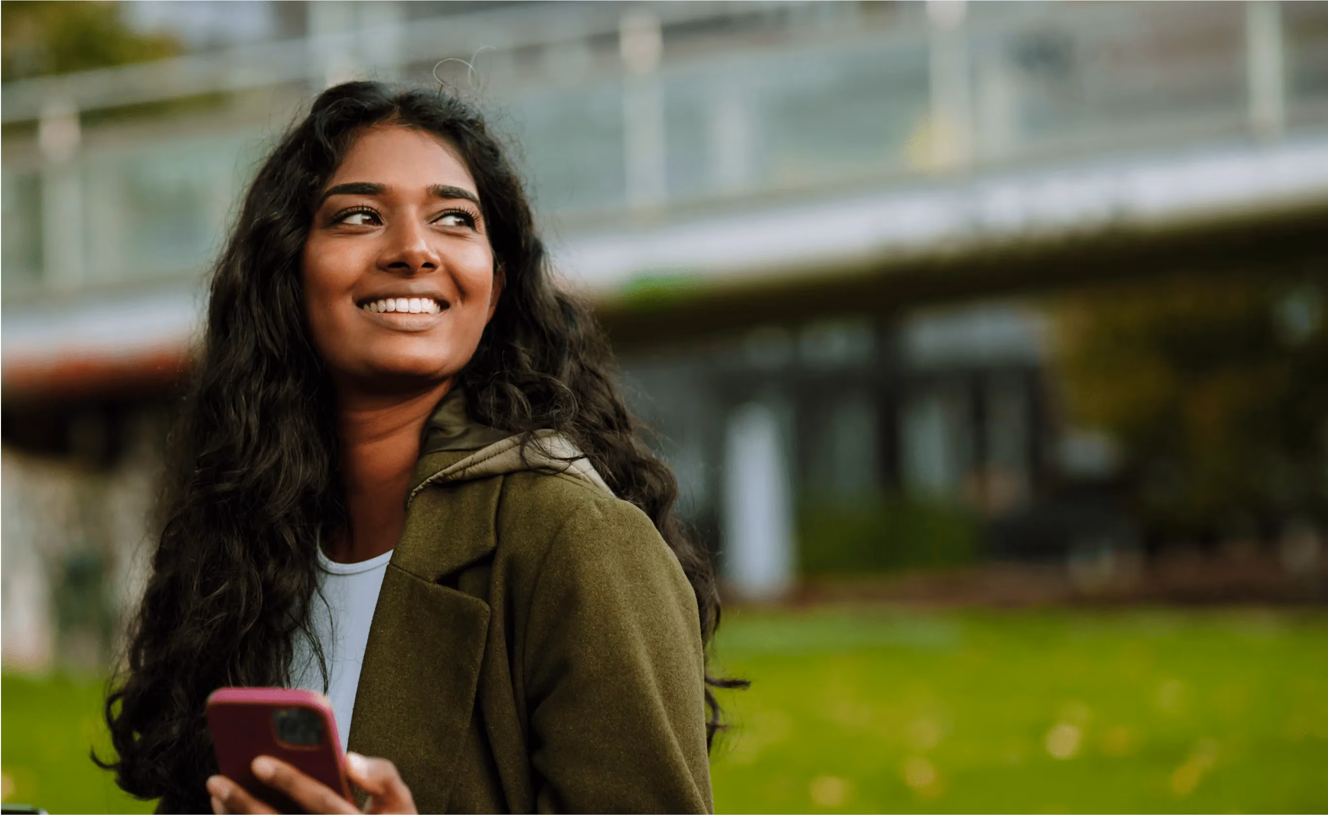 Smiling woman with long curly hair holding a smartphone outdoors with blurred green and building background.
