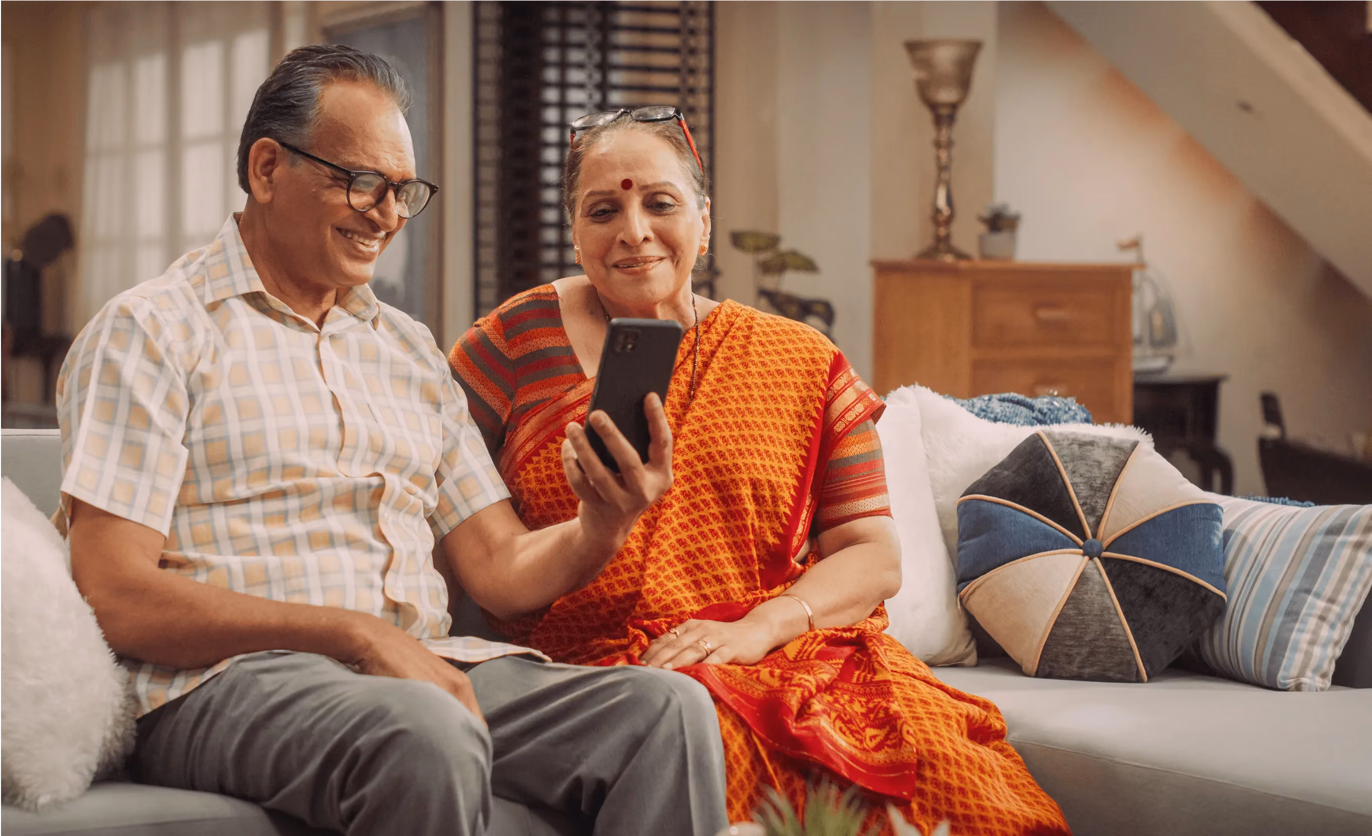 Elderly Indian couple sitting on a couch, smiling and looking at a smartphone together.