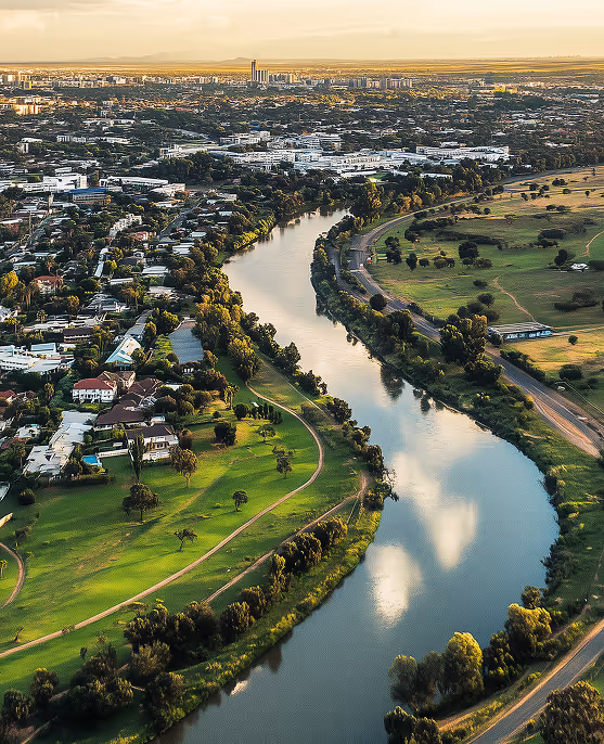 A winding river flowing through a city with residential houses on one side and green open fields on the other during golden hour.