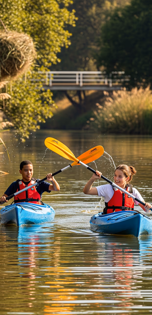 Two people in blue kayaks crossing yellow paddles on a calm river with greenery and a bridge in the background.