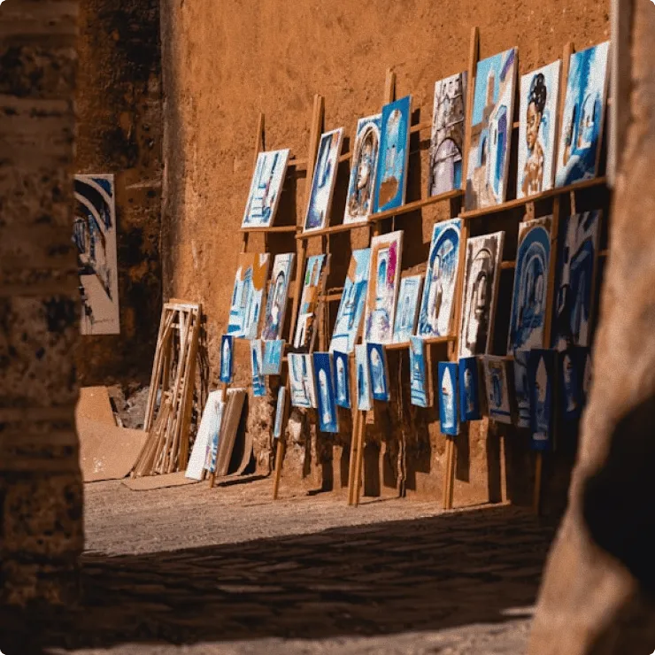 Art paintings with blue tones displayed on wooden easels against a rough textured wall outdoors.
