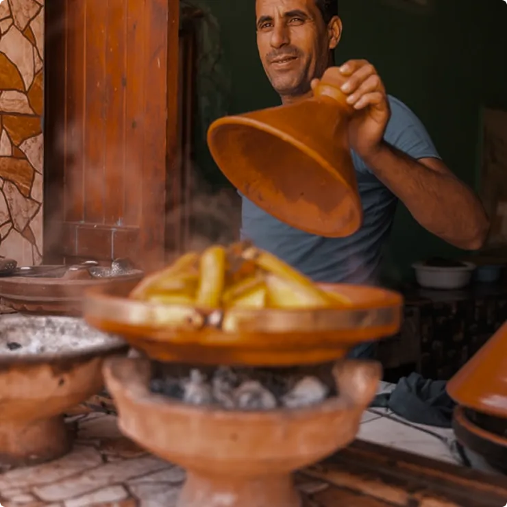 Man lifting lid of a steaming tagine cooking dish with food inside over charcoal heat.