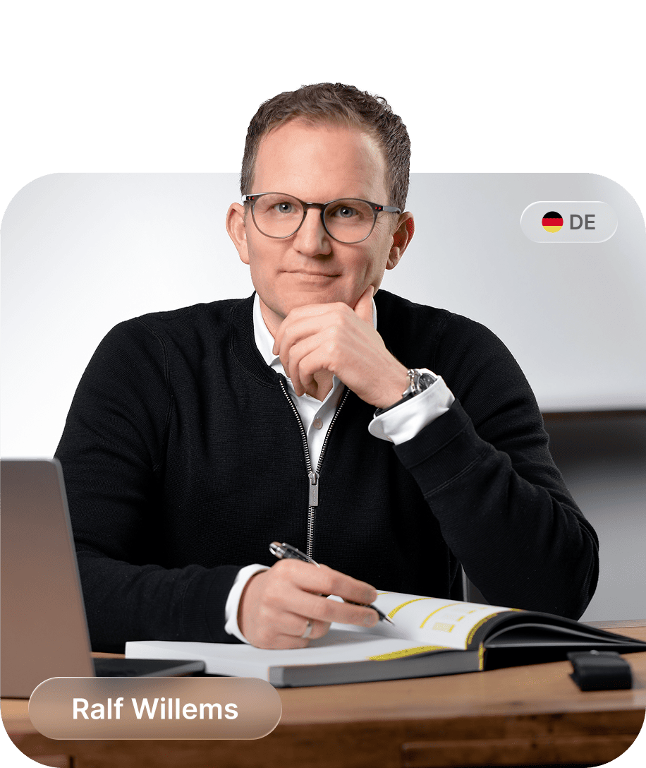 Mann mit Brille und schwarzem Pullover sitzt an einem Schreibtisch, hält einen Stift und sieht freundlich in die Kamera.