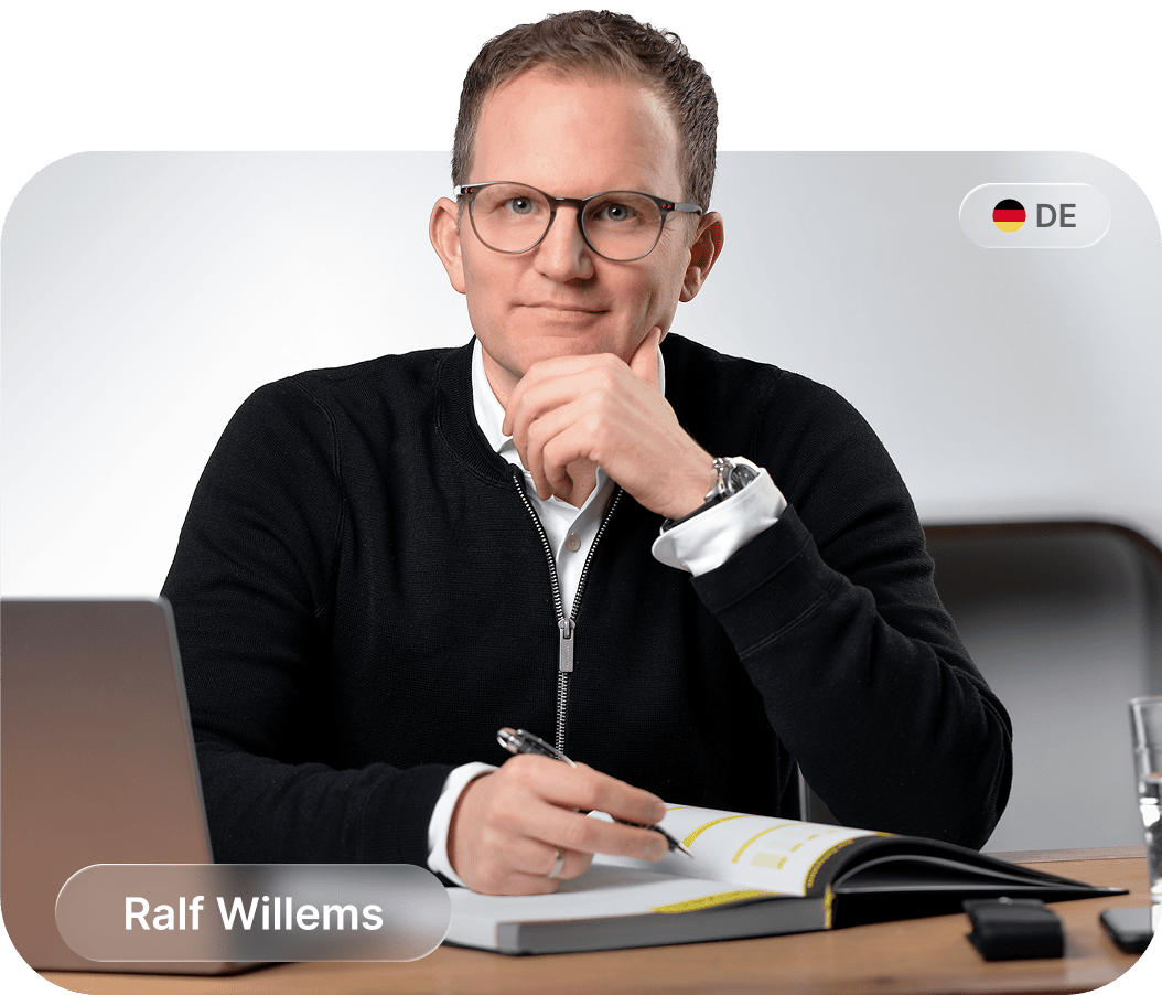 Ein Mann mit Brille und schwarzem Pullover sitzt an einem Holztisch, hält einen Stift und schaut in die Kamera.