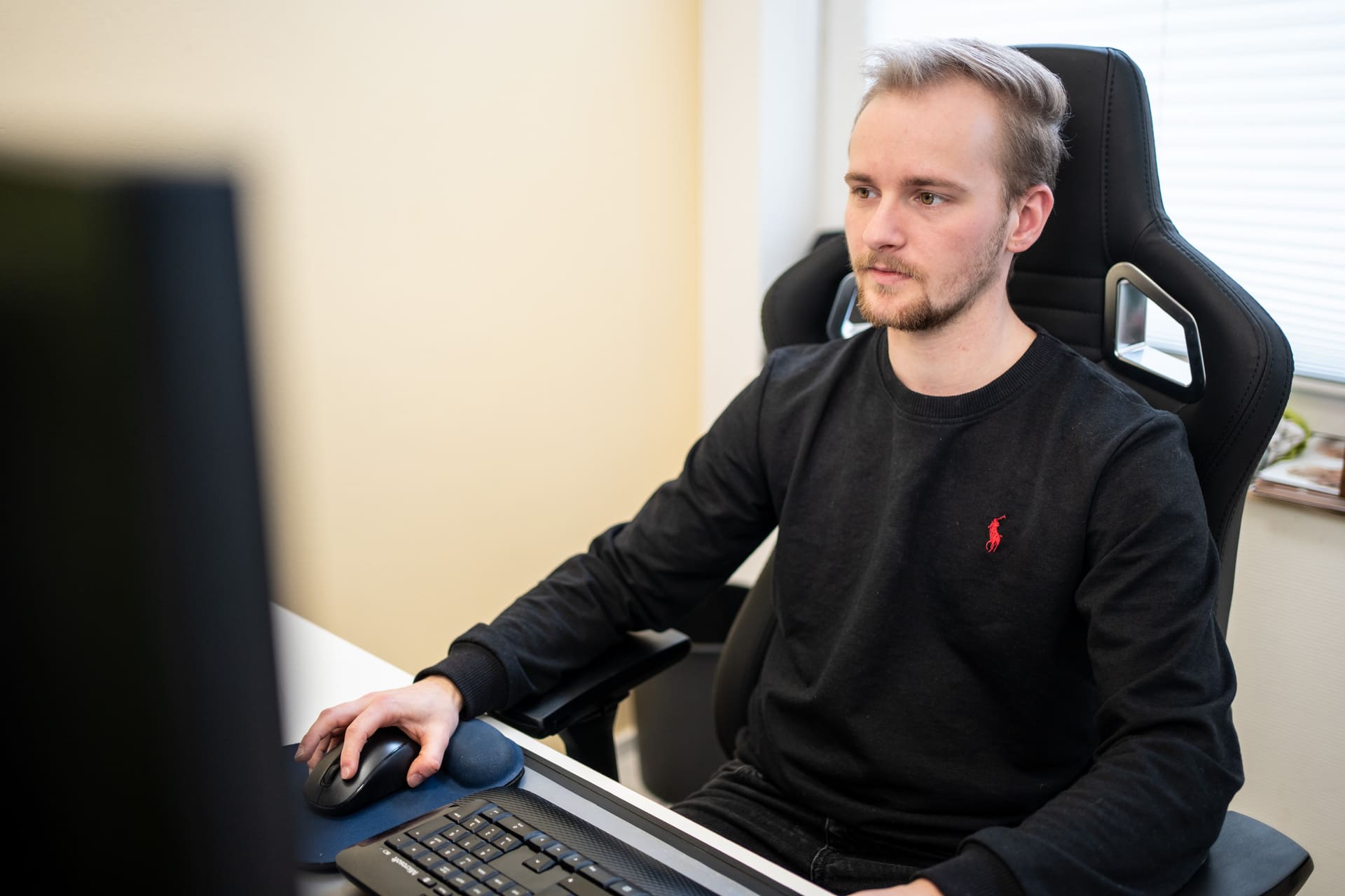 Mann mit schwarzem Pullover sitzt in ergonomischem Bürostuhl und bedient Computermaus vor Tastatur und Monitor.