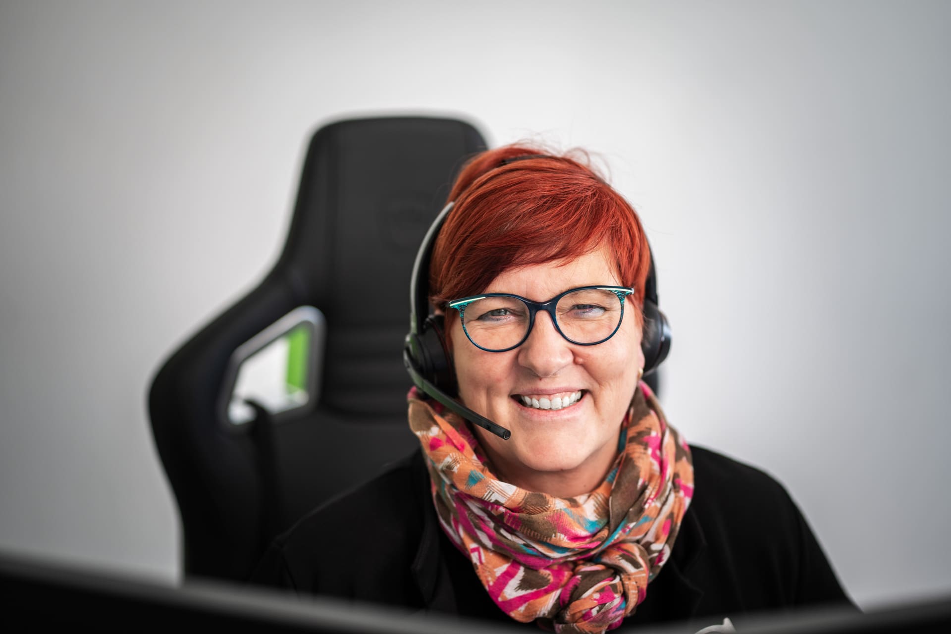Lächelnde Frau mit rotem Haar, Brille und Headset sitzt vor einem Computer, trägt bunten Schal.