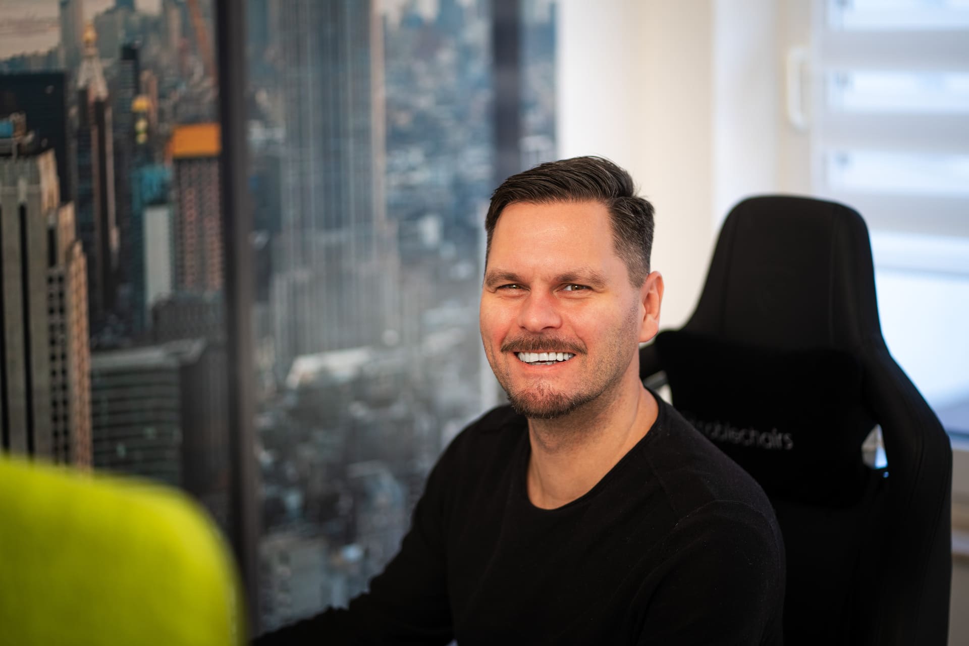 Lächelnder Mann mit kurzem braunem Haar und Bart vor einer Fensterkulisse mit Hochhausblick, sitzend auf einem schwarzen Bürostuhl.
