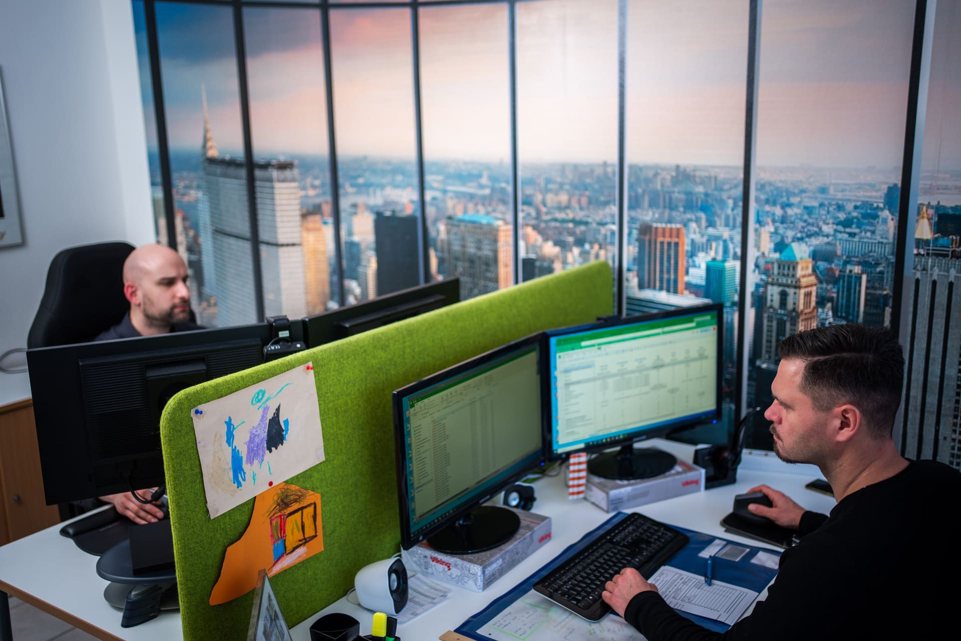 Zwei Männer arbeiten an Computern an benachbarten Schreibtischen mit einer grünen Trennwand, im Hintergrund eine Wand mit Stadtpanorama.