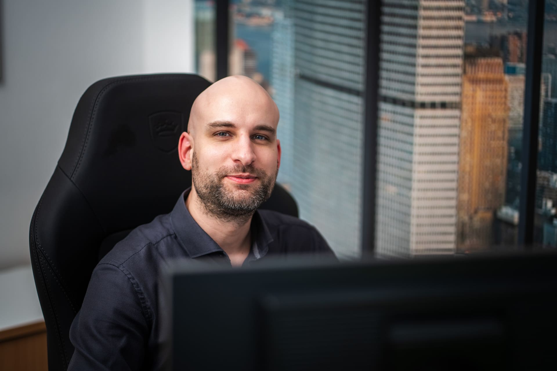 Ein lächelnder Mann mit Glatze und kurzen Bart sitzt an einem Schreibtisch vor einem Computer, im Hintergrund große Fenster mit Blick auf Wolkenkratzer.