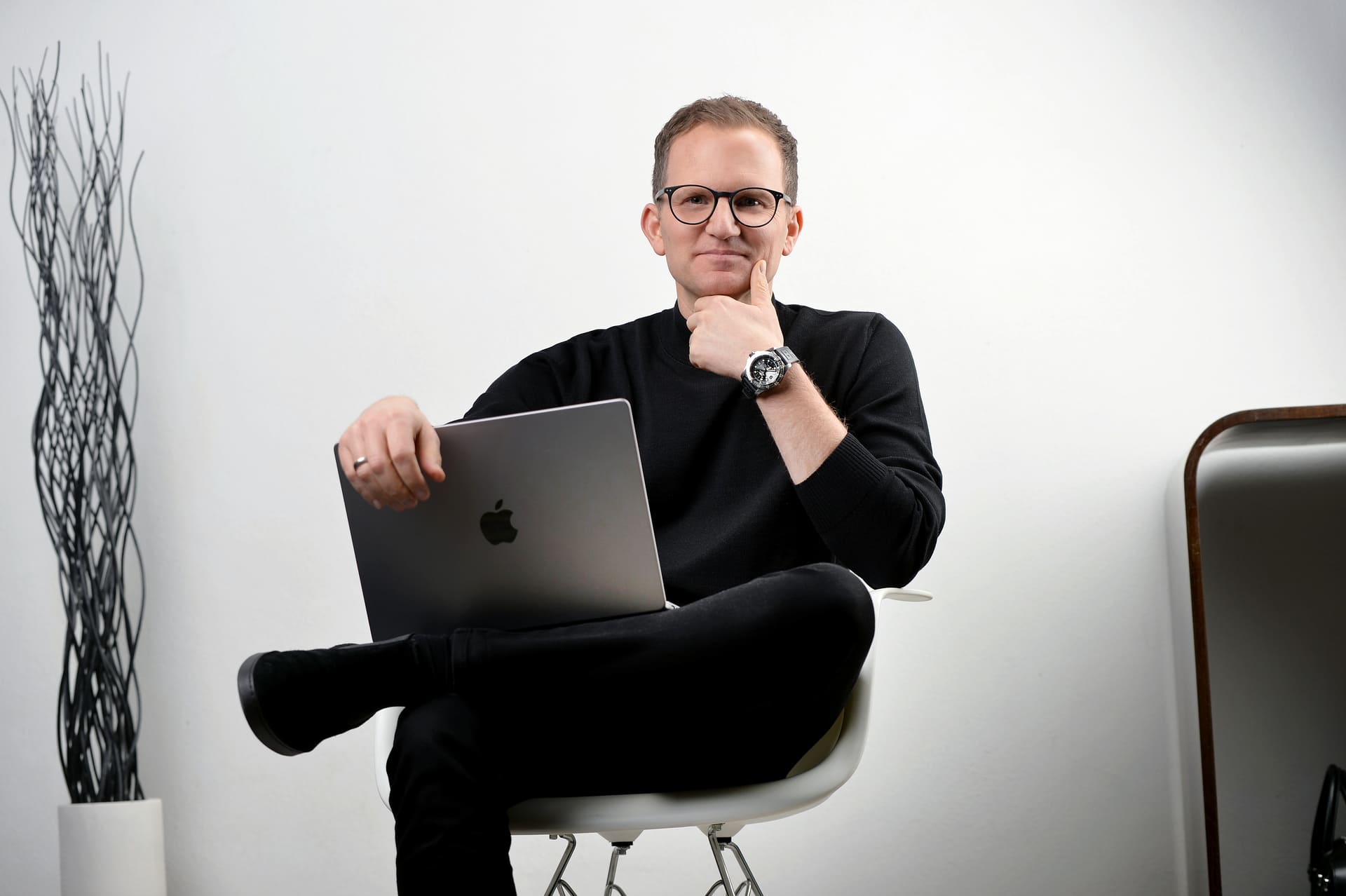 Mann mit Brille und schwarzem Pullover sitzt mit verschränkten Beinen auf einem weißen Stuhl und hält einen Laptop auf dem Schoß.