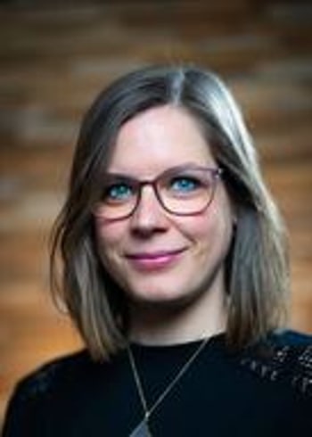 Portrait einer Frau mit mittellangen braunen Haaren, die eine Brille trägt und in die Kamera lächelt, vor einem verschwommenen Hintergrund aus Ziegelsteinen.