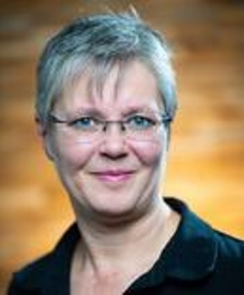 Porträt einer lächelnden Frau mit kurzen grauen Haaren und Brille vor unscharfem orangefarbenem Hintergrund.