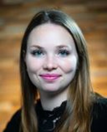 Porträt einer jungen Frau mit langen braunen Haaren, die in die Kamera lächelt, vor einem verschwommenen, warmen Hintergrund.