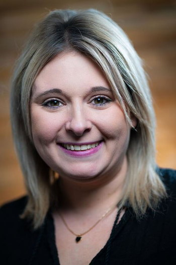 Porträt einer lächelnden Frau mit blonden Haaren und schwarzer Oberbekleidung vor unscharfem braunem Hintergrund.