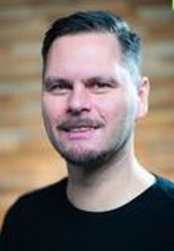 Porträt eines lächelnden Mannes mit kurzem dunklem Haar und kurzem Bart vor unscharfem braunem Hintergrund.