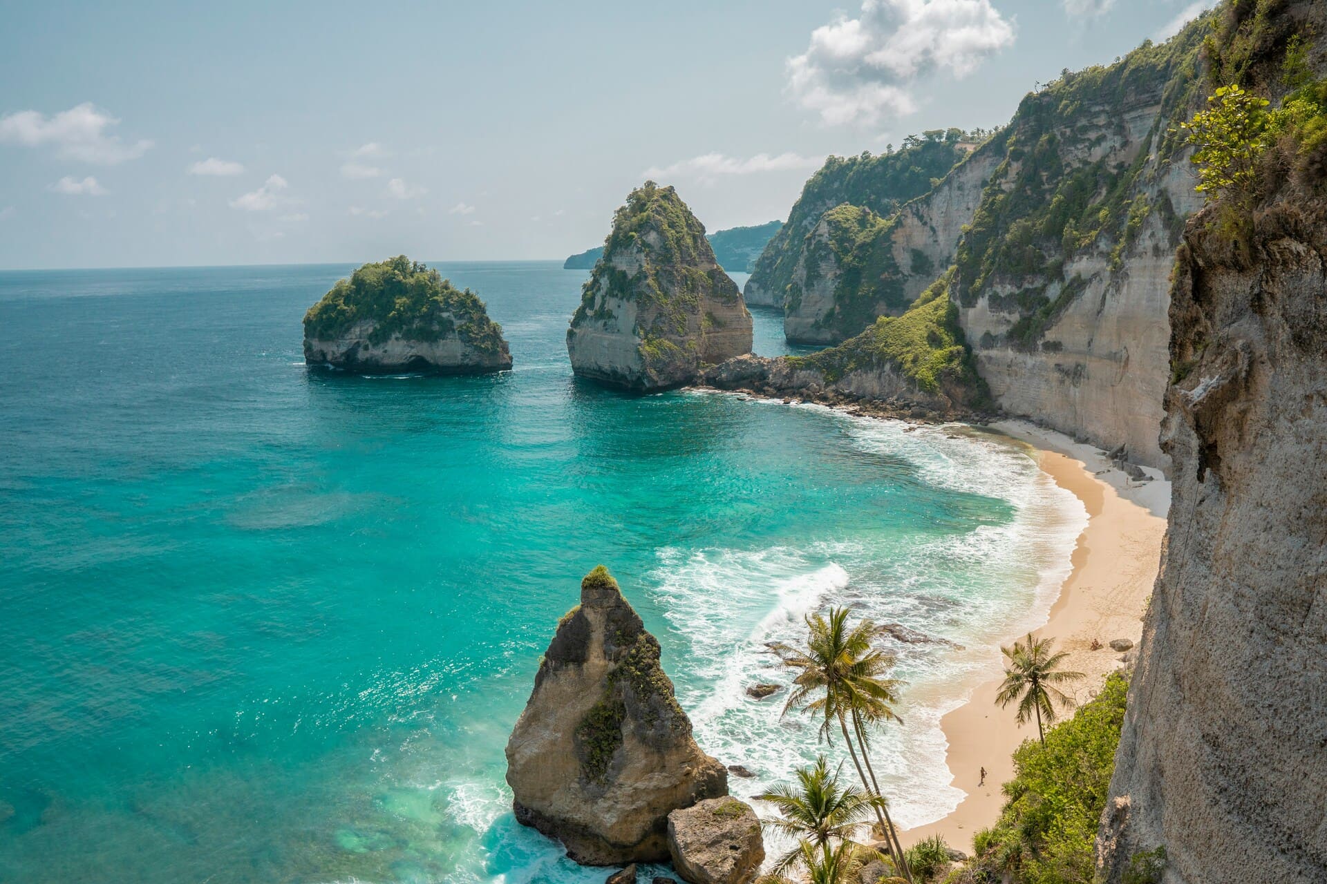 Türkisfarbenes Meer mit felsigen Inseln und einem Sandstrand umgeben von bewaldeten Klippen unter einem bewölkten Himmel.