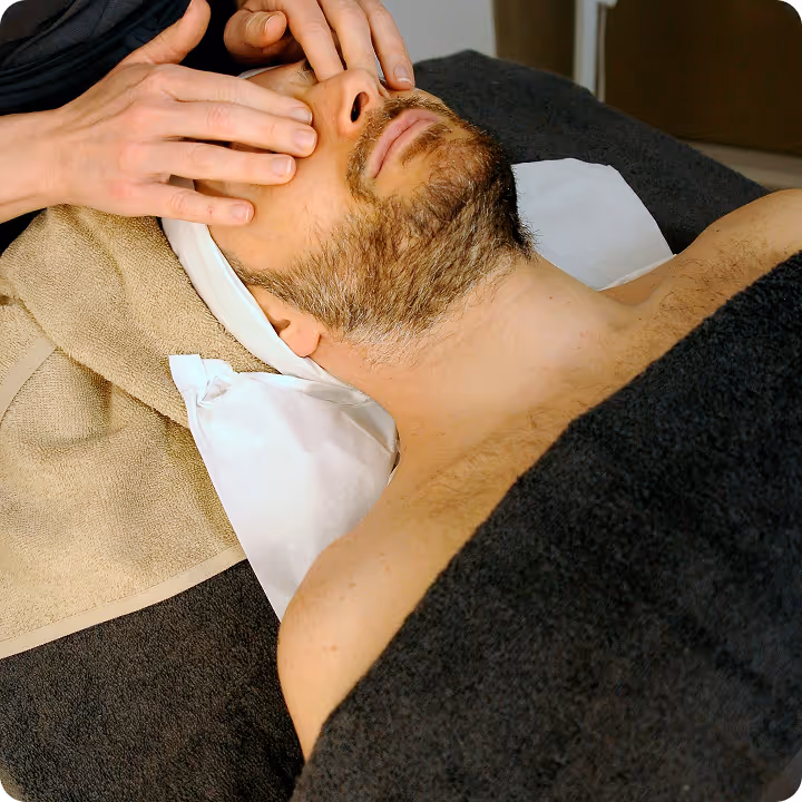 Man with a beard receiving a facial massage while lying on a spa bed covered with a dark towel.