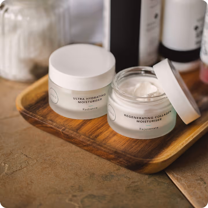 Two skincare moisturizer jars on a wooden tray, one closed labeled Ultra Hydrating Moisturiser, the other open labeled Regenerating Collagen Moisturiser with cream inside.
