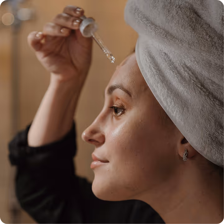 Woman with a towel wrapped around her head applying skincare serum to her face using a dropper.