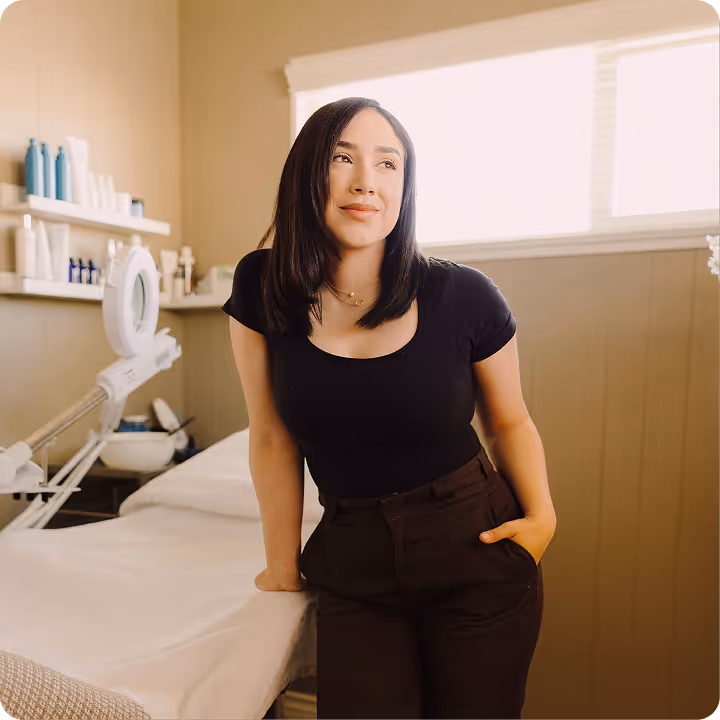 Woman with shoulder-length dark hair leaning against a spa treatment bed in a softly lit room.