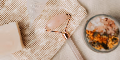 Rose quartz facial roller placed on woven fabric with a bowl of dried flowers and a white soap bar nearby.