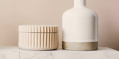 Beige ceramic jar with vertical ridges next to a tall white vase with a beige base on a white surface.