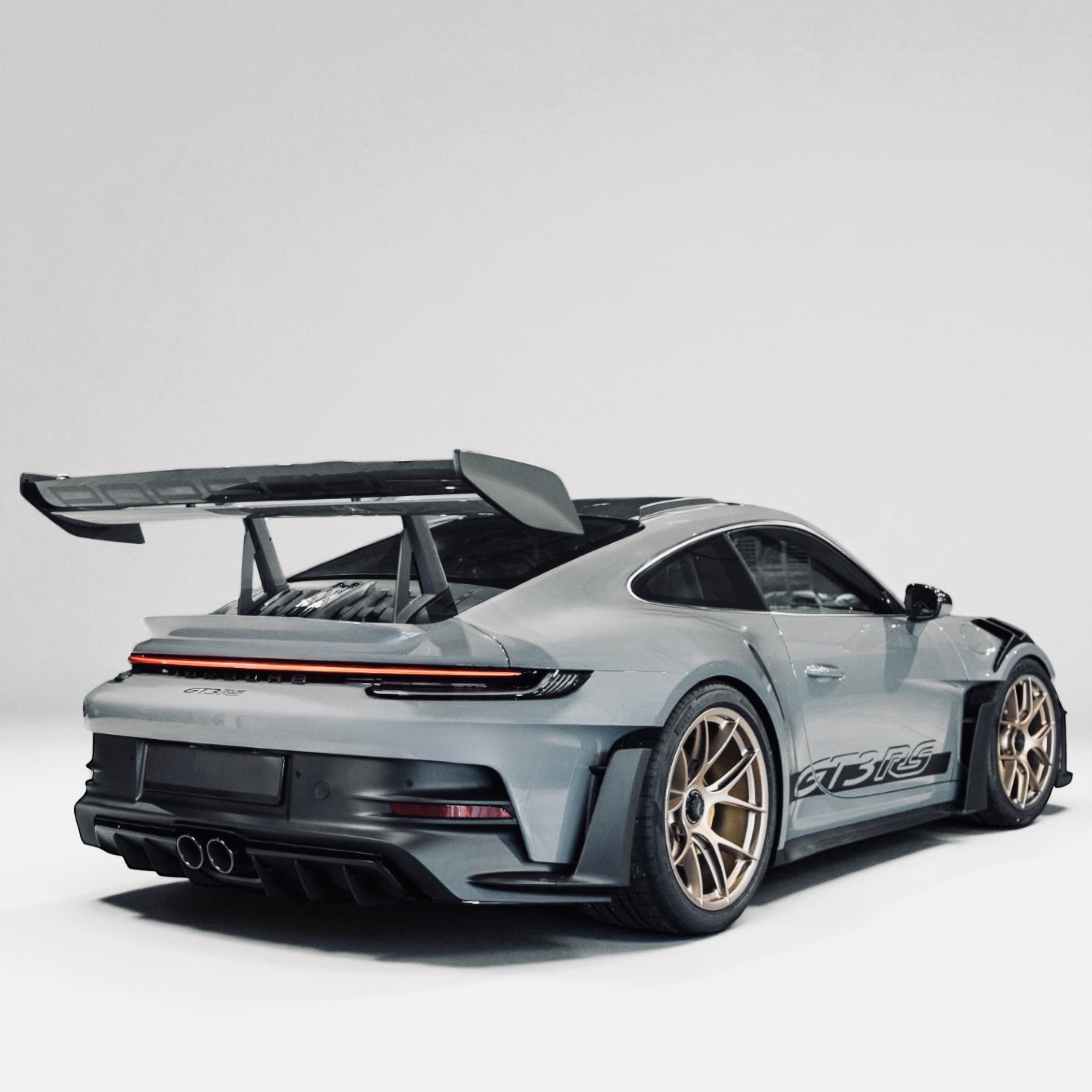 Rear side view of a gray Porsche 911 GT3 RS with a large rear spoiler and bronze wheels on a white background.