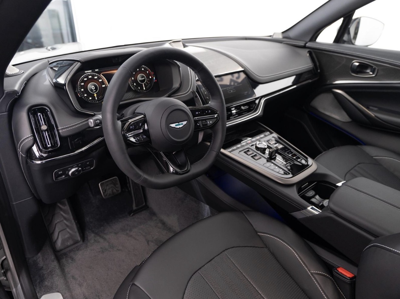 Interior of a luxury car showing a black leather steering wheel with Aston Martin logo, digital dashboard, and center console with controls and gear selector.