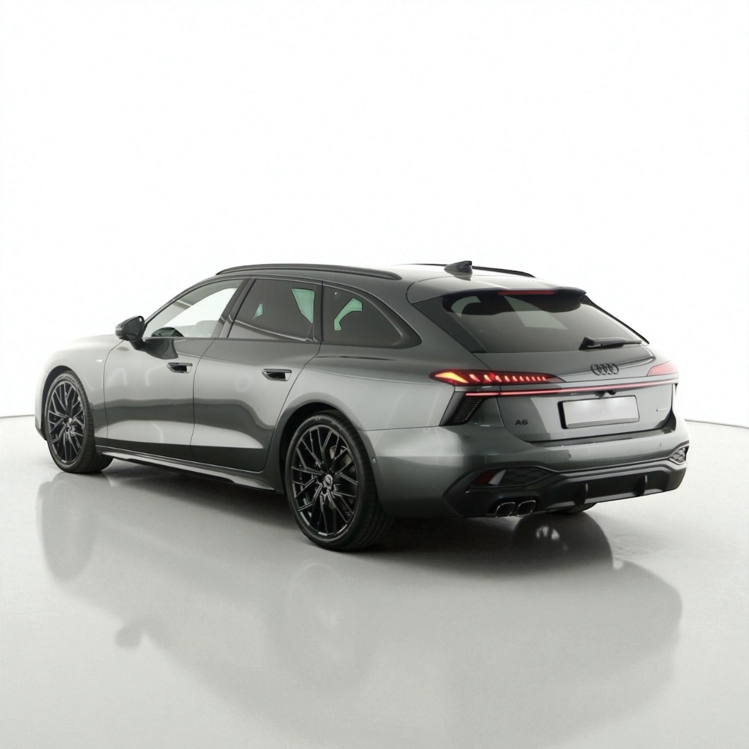 Rear side view of a gray Audi A6 Avant station wagon with black rims and illuminated rear lights on a white reflective floor.