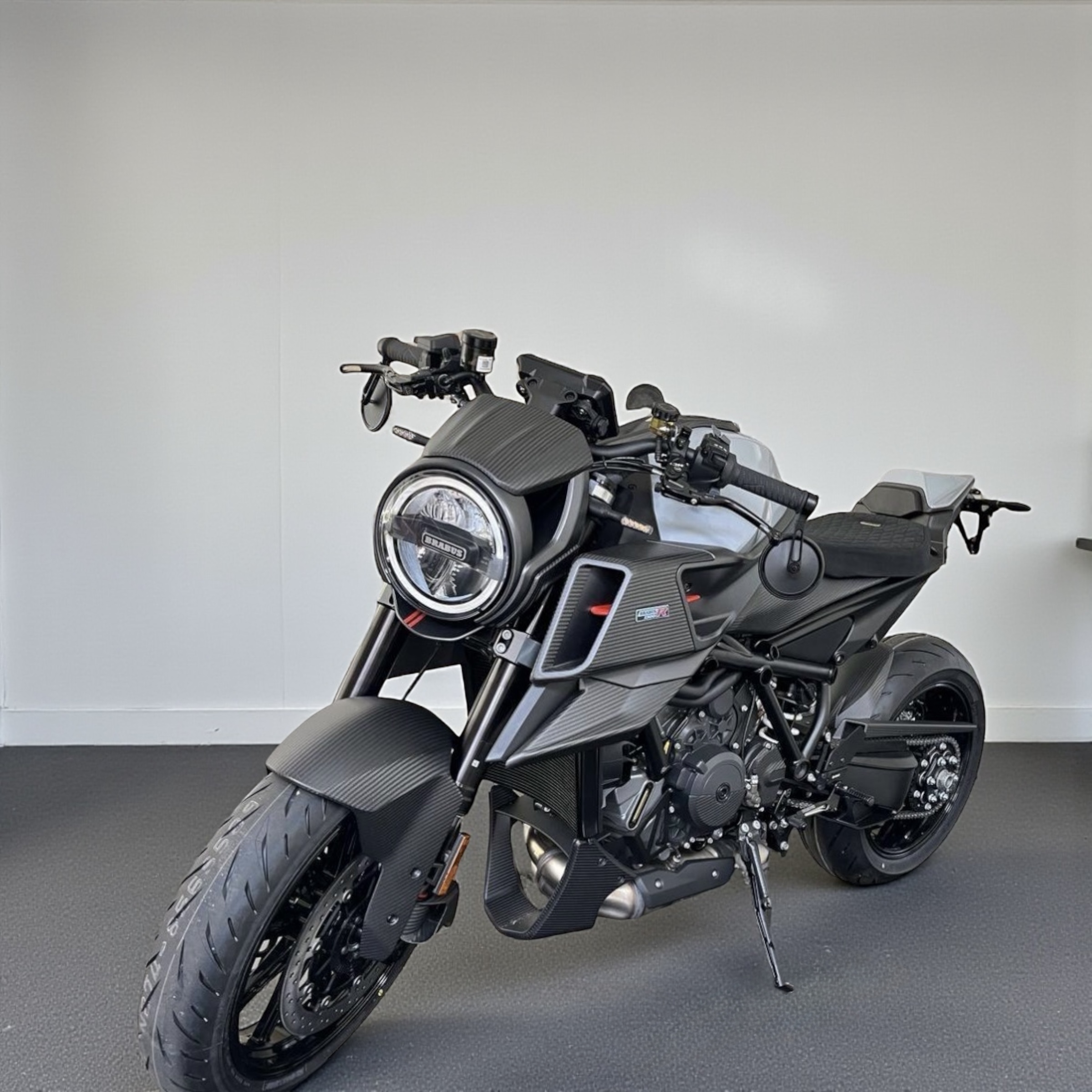 Sleek black modern motorcycle with round headlight and carbon fiber detailing parked indoors on gray flooring.