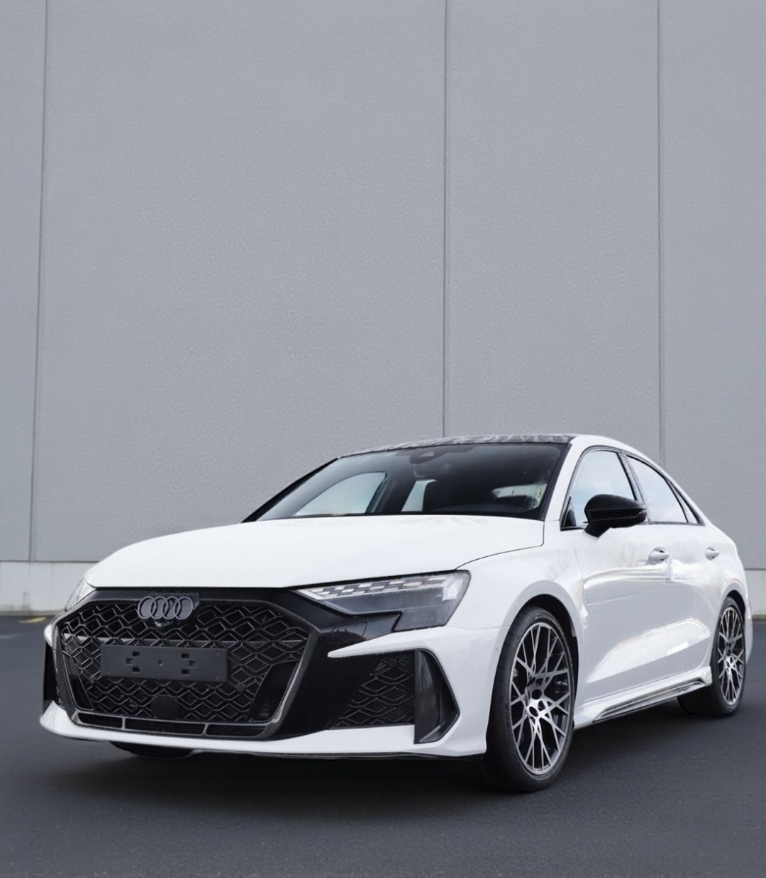 White Audi sedan parked on a dark pavement in front of a plain gray wall.