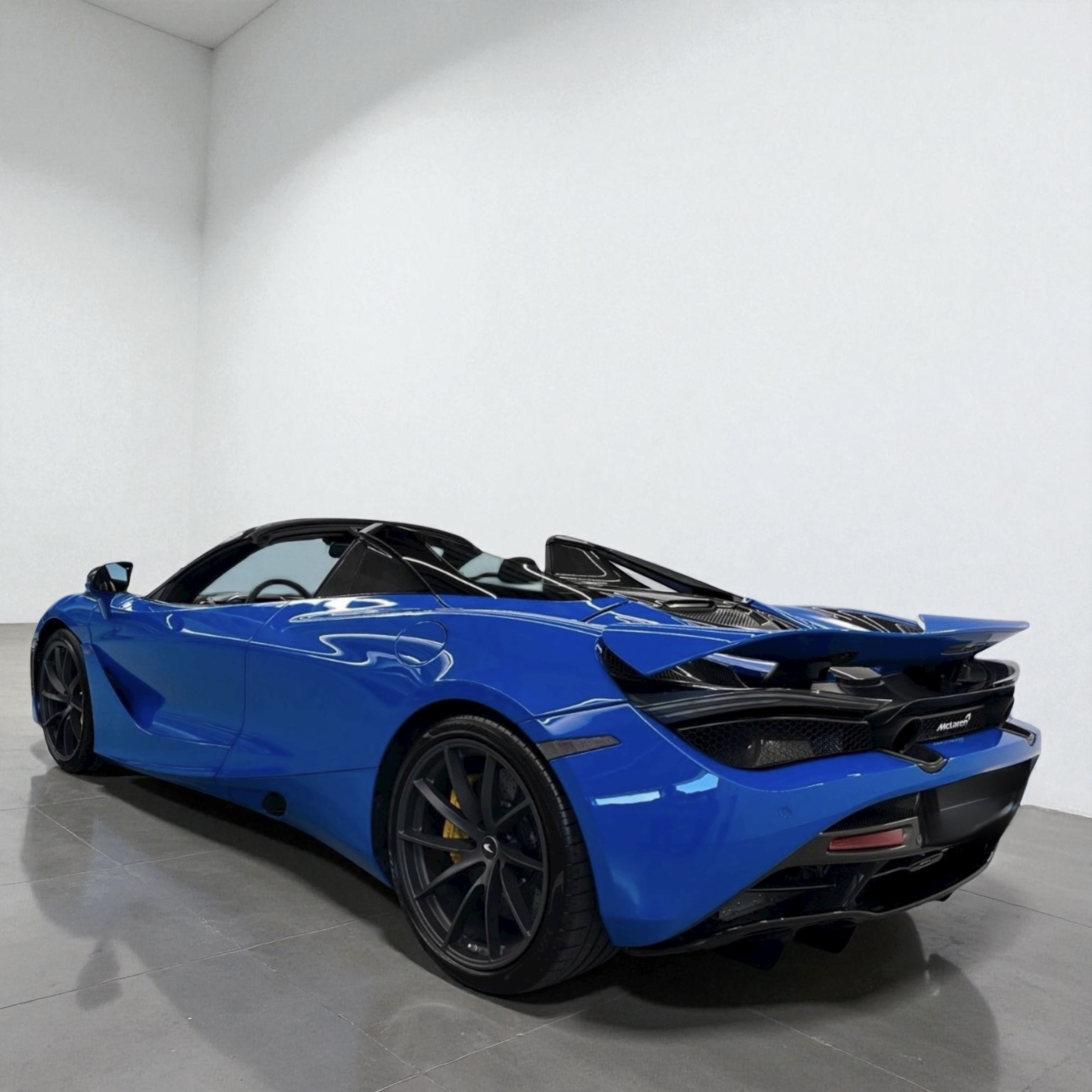 Blue McLaren sports car with black roof and rear spoiler parked indoors on gray floor.