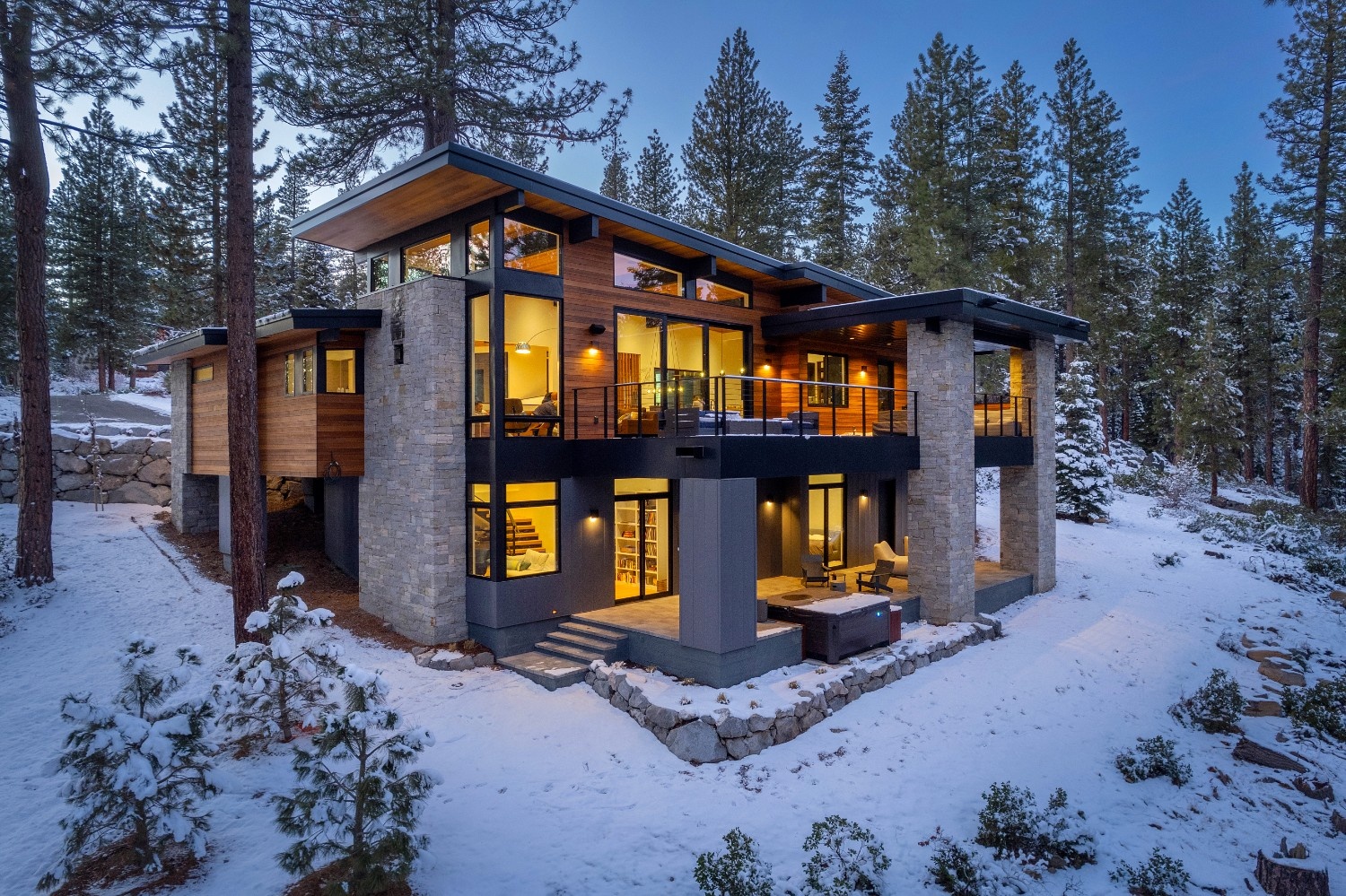 Modern two-story house with large windows and warm interior lighting surrounded by snow-covered ground and pine trees at dusk.