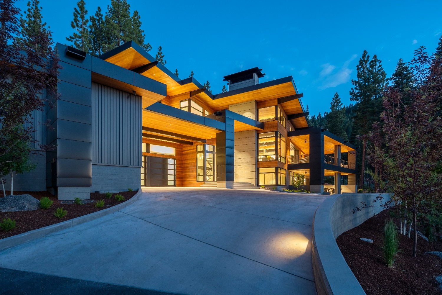 Modern two-story house with warm exterior lighting, large windows, and concrete driveway surrounded by trees at dusk.