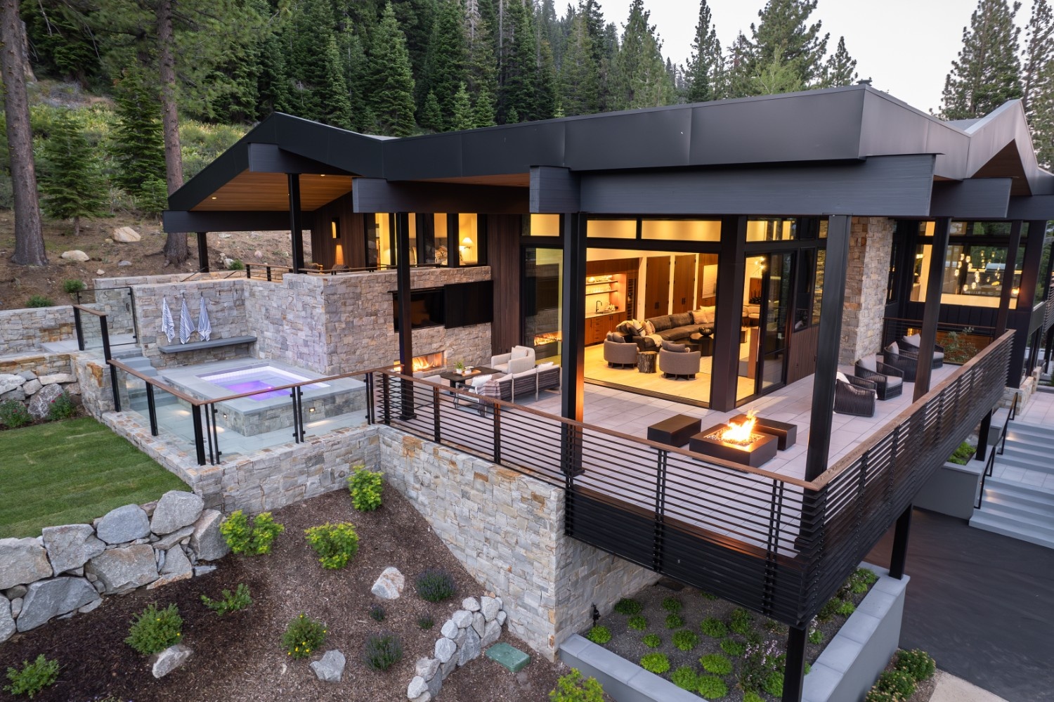 Modern mountain home with illuminated living room, outdoor fireplace, and a hot tub surrounded by stone walls and pine trees.
