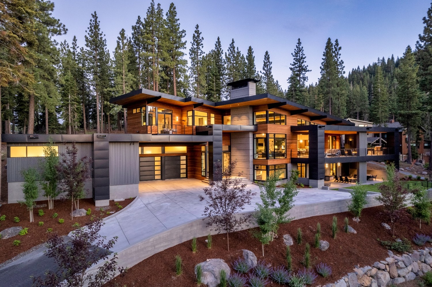 Modern large house with wood and stone exterior surrounded by tall pine trees at dusk.