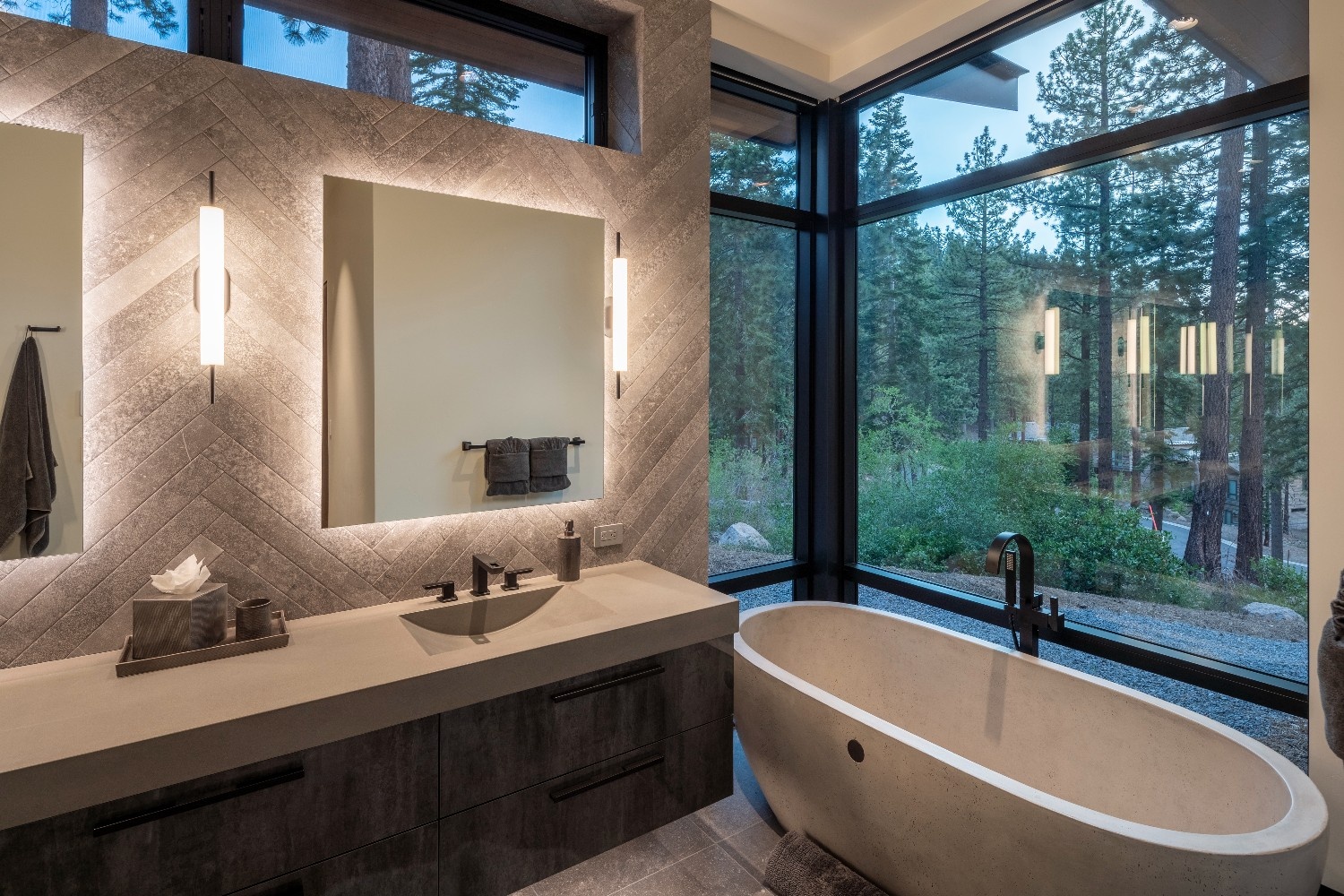 Modern bathroom with concrete bathtub by a large corner window showing forest view and a vanity with illuminated mirror and wall sconces.