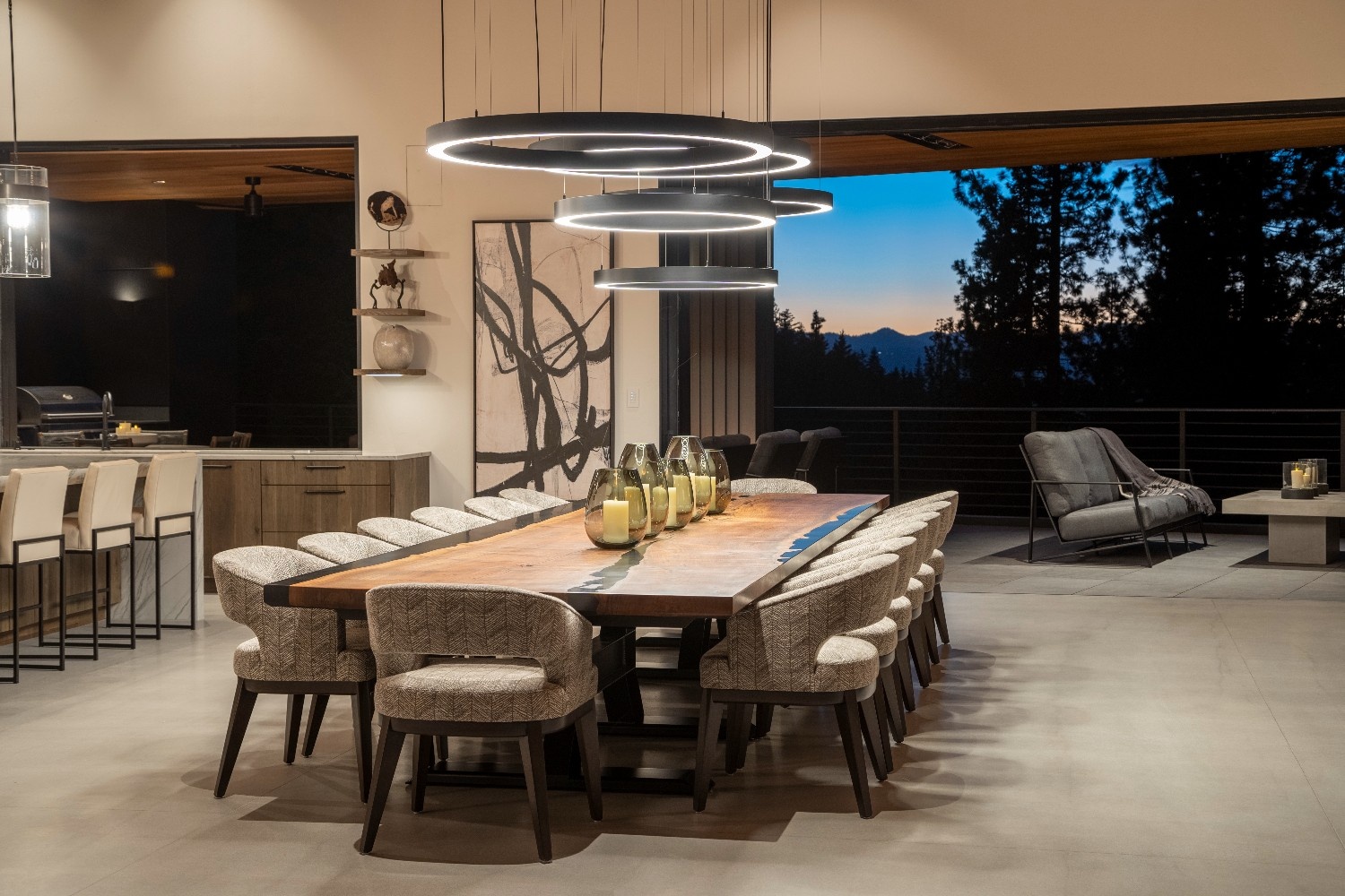 Modern dining room with a long wooden table surrounded by upholstered chairs, circular LED pendant lights, and a large window showing a twilight forest view.