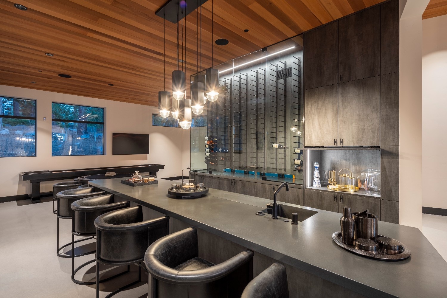 Modern home bar with black leather stools, dark countertop, pendant lights, glass wine rack, and wooden ceiling.