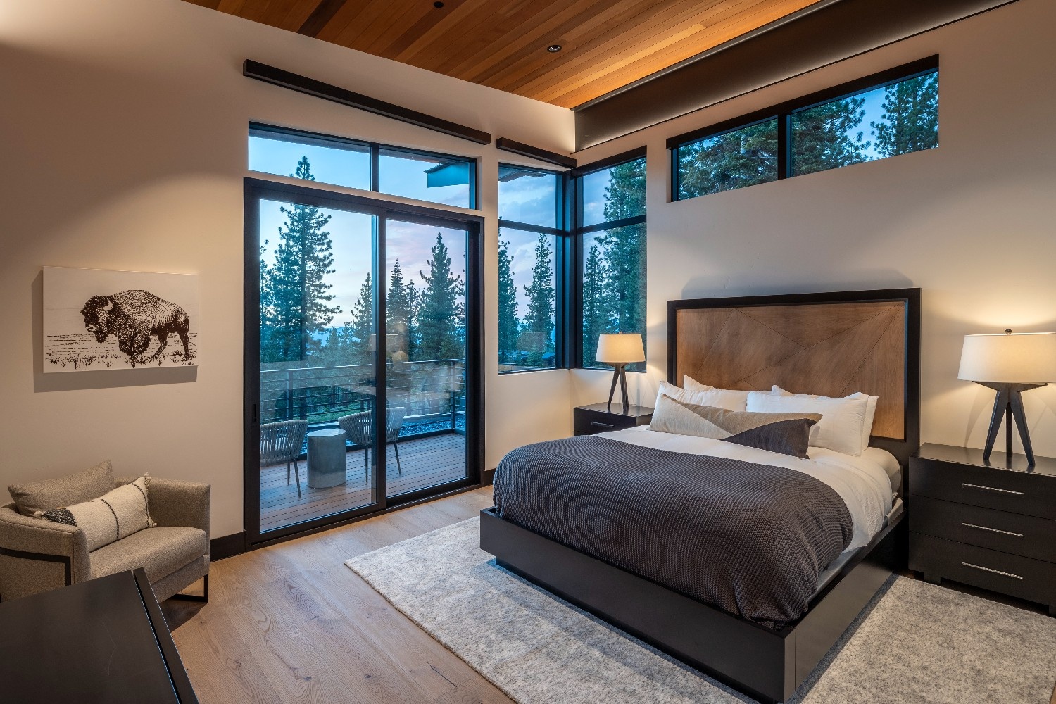 Modern bedroom with large windows showing a forest view, a bed with wooden headboard, and two bedside lamps.