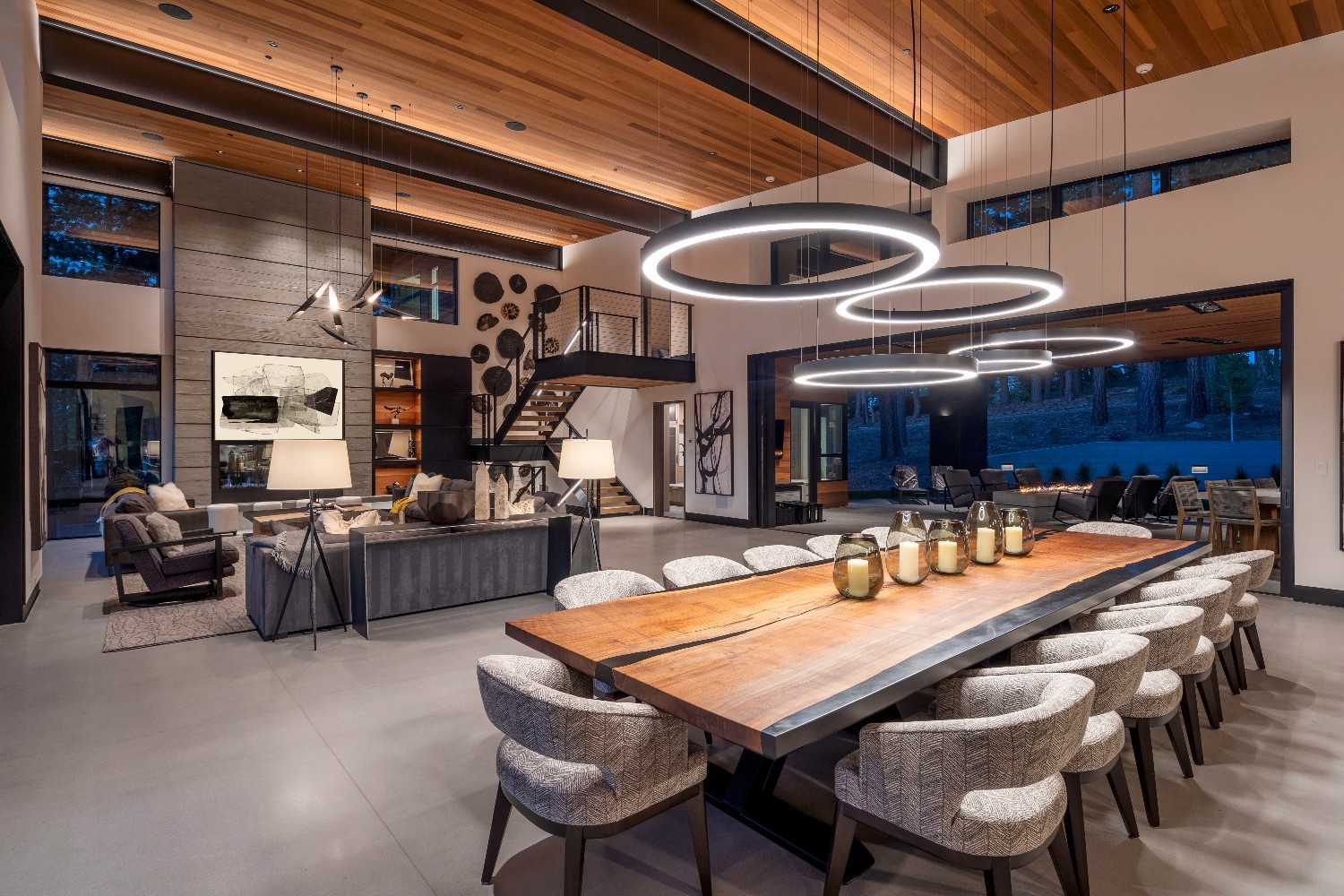 Modern spacious dining and living area with a long wooden table surrounded by upholstered chairs, circular pendant lights, and large sliding doors opening to an outdoor seating area.