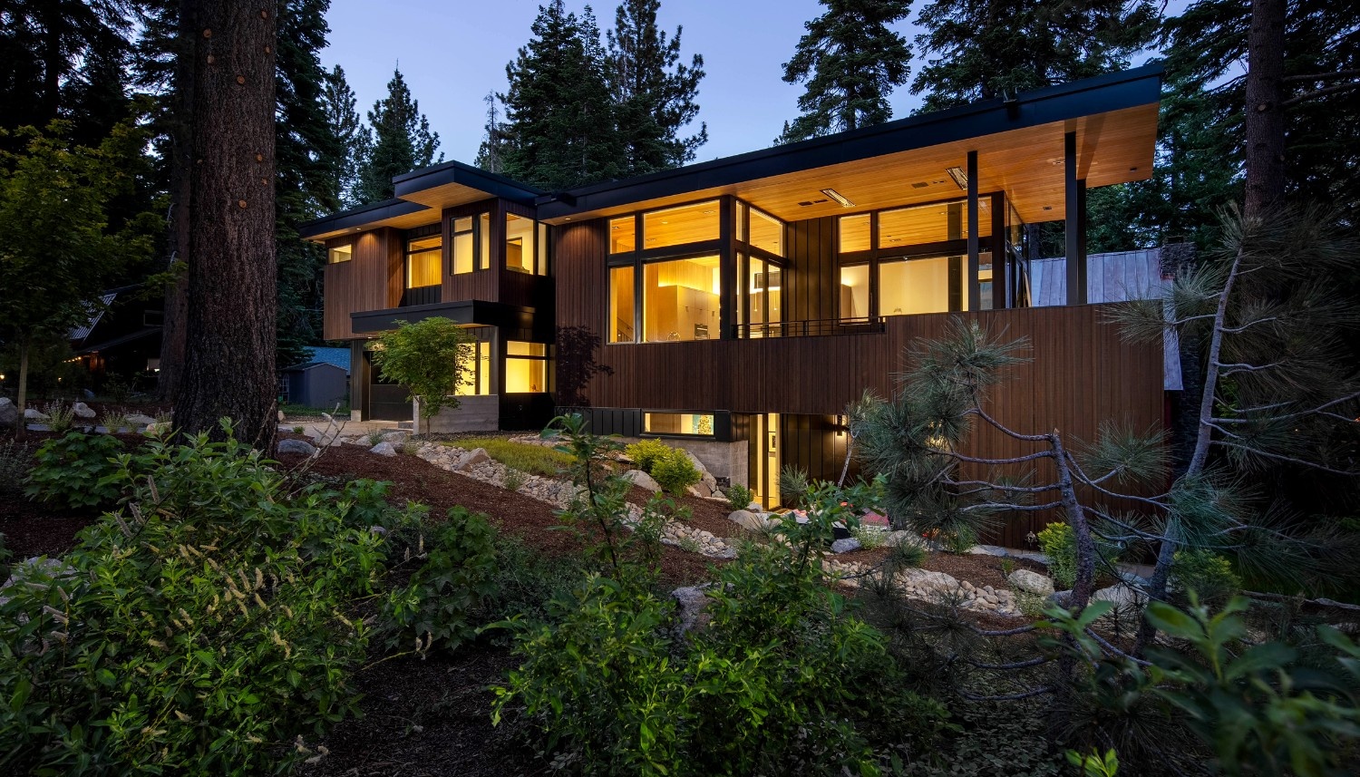 Modern two-story wooden house with large windows illuminated at dusk, surrounded by trees and greenery.
