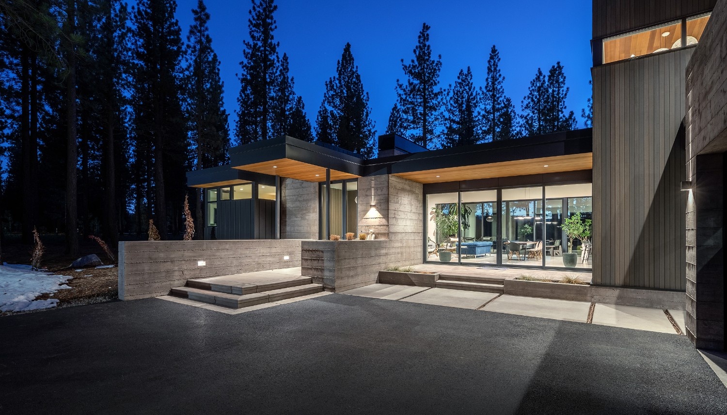 Modern house exterior at dusk with large glass windows, concrete walls, wood-paneled overhangs, and surrounding tall pine trees.