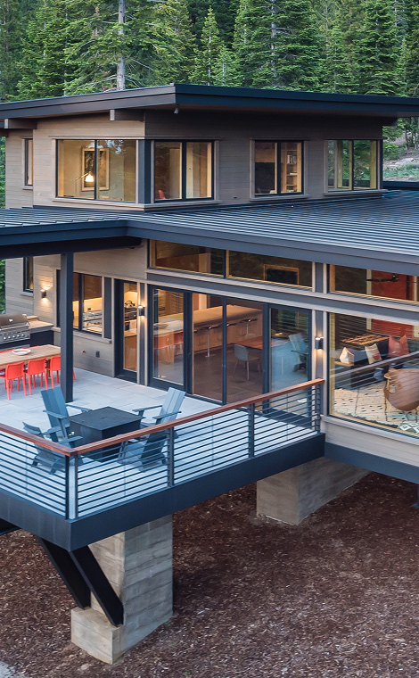Modern two-story house with large windows, a balcony with outdoor chairs and a fire pit, surrounded by dense pine trees.