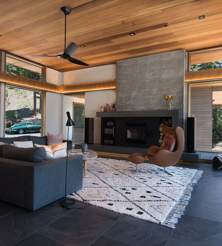 Modern living room with wooden ceiling, large windows, gray sofa, brown leather armchair, patterned white rug, and a concrete fireplace.