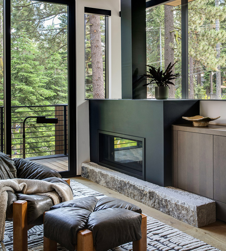 Modern living room corner with black leather armchair and ottoman, stone hearth fireplace, large windows showing green trees outside.