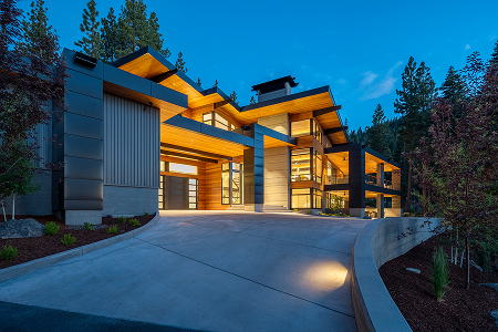 Modern two-story house with large windows and warm interior lighting at dusk, surrounded by trees.