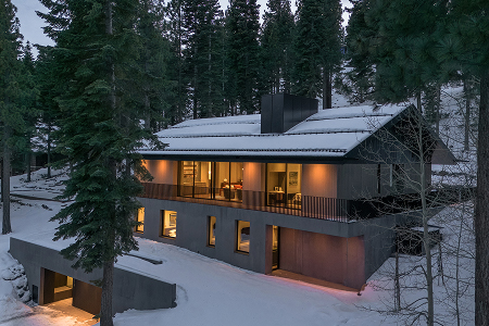Modern two-story house with large windows and warm interior lights surrounded by snow-covered ground and tall pine trees at dusk.